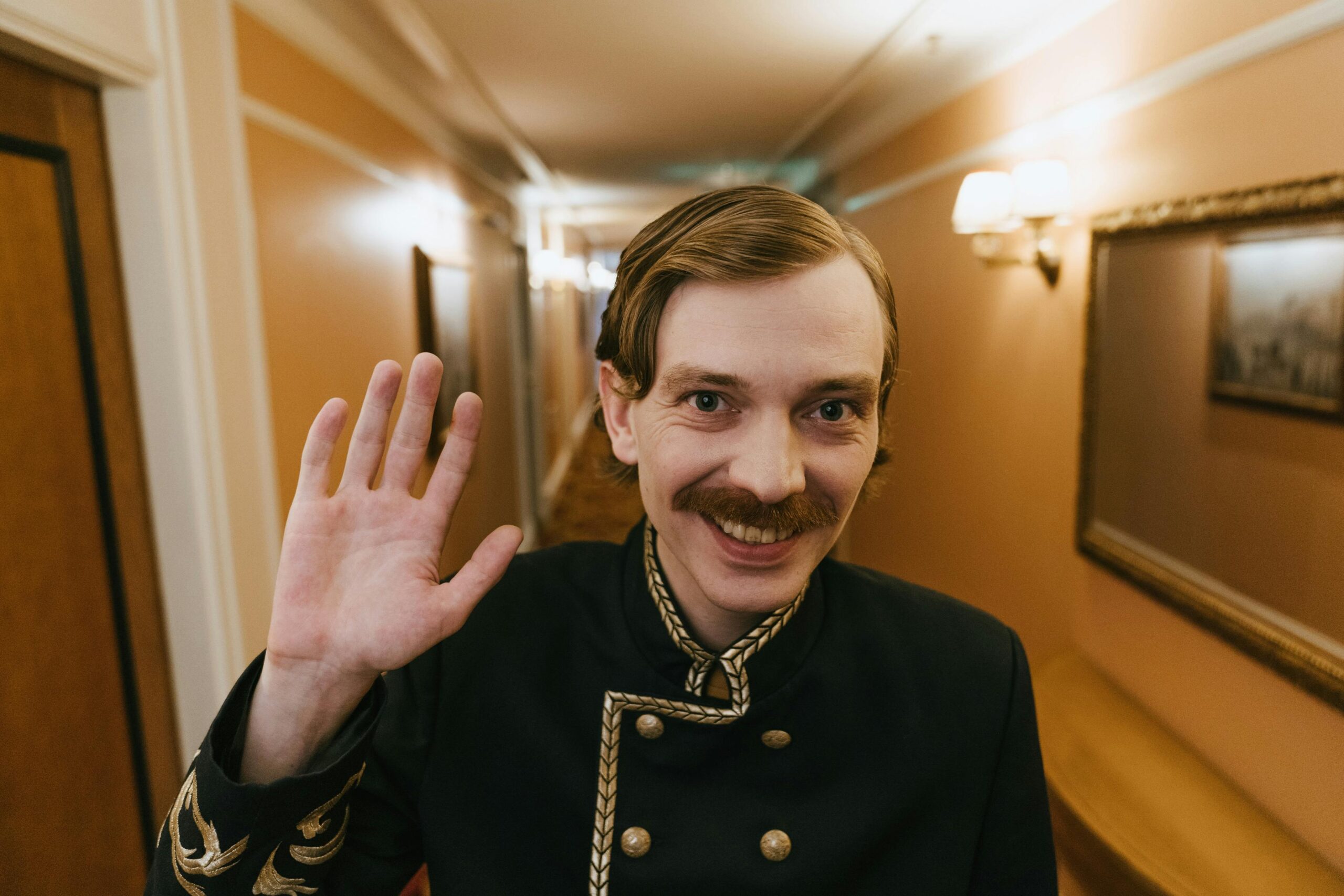 A smiling bellboy with a moustache waves in a hotel hallway, creating a welcoming atmosphere.